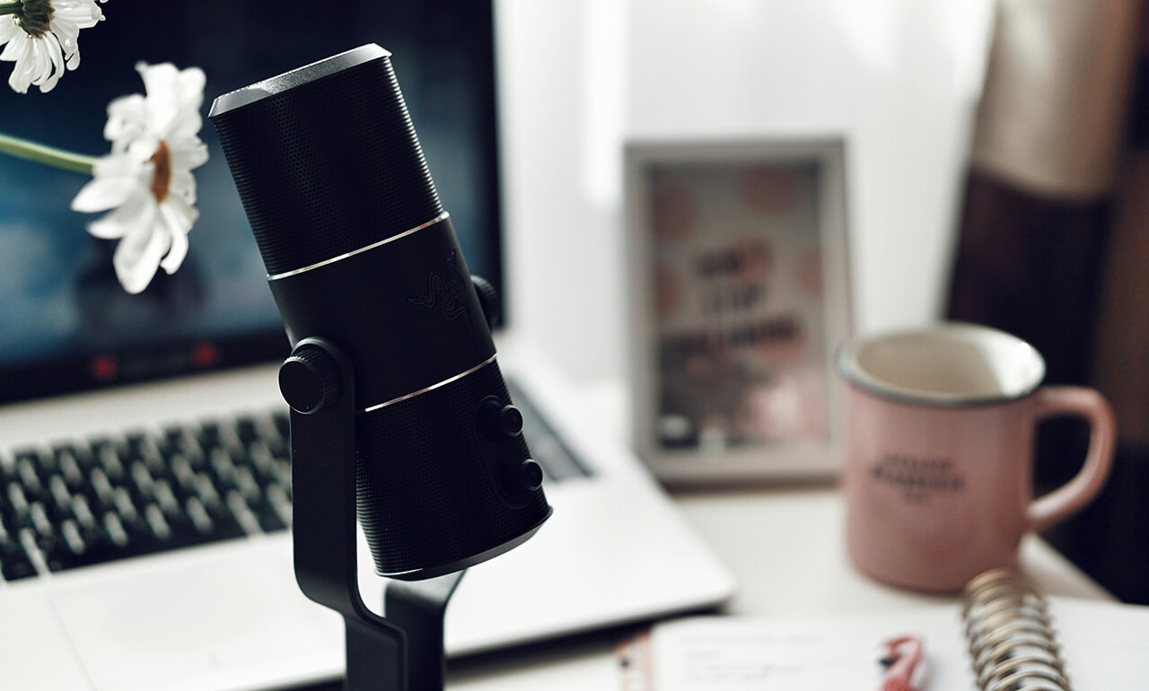 Ein Schreibtisch mit einem Mikrofon im Vordergrund und Laptop, Kaffeetasse und Notizblock im Hintergrund.